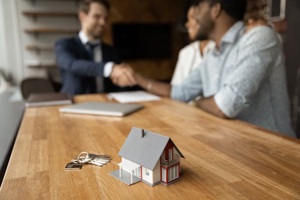 Using a 1031 exchange on a primary residence is illustrated by a couple shaking hands with a real estate agent while closing the sale of their house.